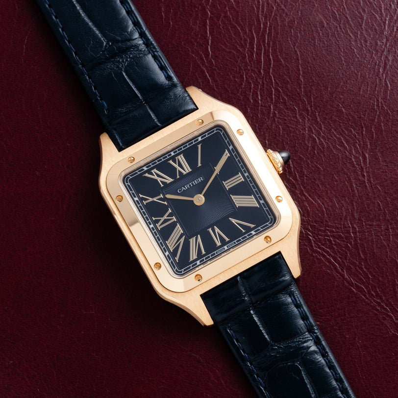 The Cartier Santos Dumont watch, featuring a black leather strap and Roman numerals, rests on a dark red textured surface.