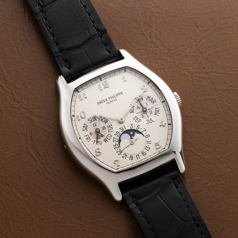 The Patek Philippe Tonneau Perpetual Calendar wristwatch features a silver case and black leather strap, displayed on a brown leather surface with multiple dials.