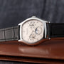 The Patek Philippe Tonneau Perpetual Calendar wristwatch with a black leather strap is displayed on a brown leather surface.