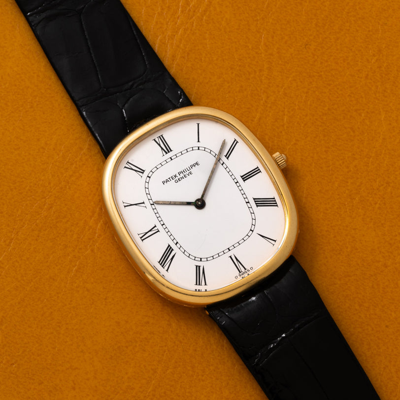 A Patek Philippe Ellipse gold watch featuring a white Roman numeral dial and black leather strap, displayed on a mustard background.