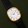 A Patek Philippe Ellipse wristwatch featuring a gold case, white dial with Roman numerals, and black leather strap displayed on a green surface.