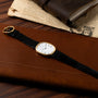 A Patek Philippe Ellipse gold wristwatch with a black leather strap rests on a brown leather surface beside a journal and newspaper.