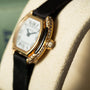 Close-up of the Cartier Ellipse, a luxury gold watch featuring diamond accents and a black leather strap, presented on a cream display cushion.