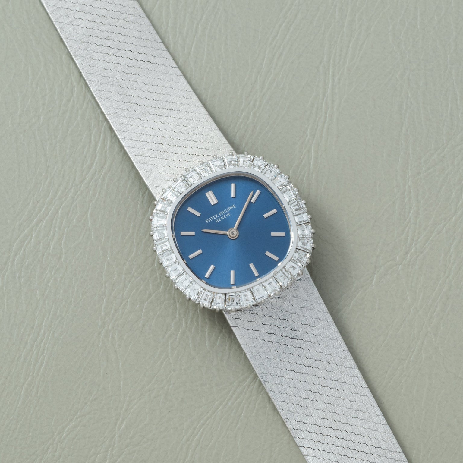 The Patek Philippe Ellipse Ladies Dress Watch features a blue face, diamond bezel, and silver strap, displayed on a light gray textured surface.