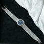 The Patek Philippe Ellipse Ladies Dress Watch, featuring a blue face and diamond bezel, is displayed on a black surface beside a white cloth.