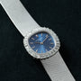 A Patek Philippe Ellipse Ladies Dress Watch in silver with a blue face and diamond bezel is displayed against a textured dark background.