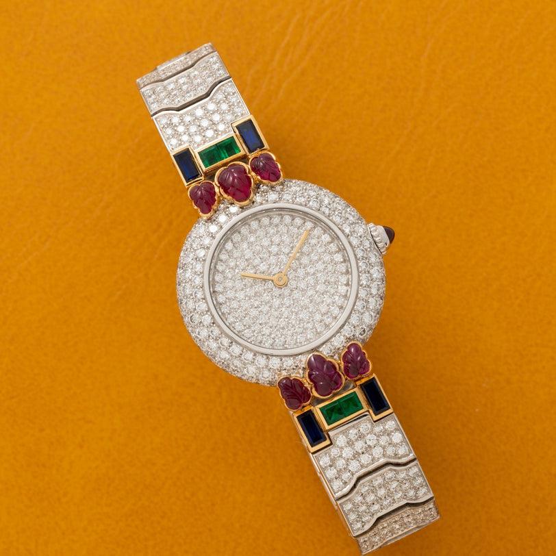 The Cartier Colisee Tutti Frutti Diamond watch features red, green, and blue gemstones on a mustard yellow background.