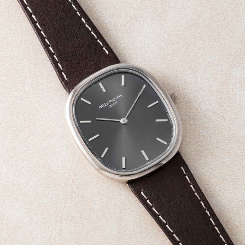 The Patek Philippe Ellipse watch features a dark dial and a brown leather strap, displayed on a light textured surface.