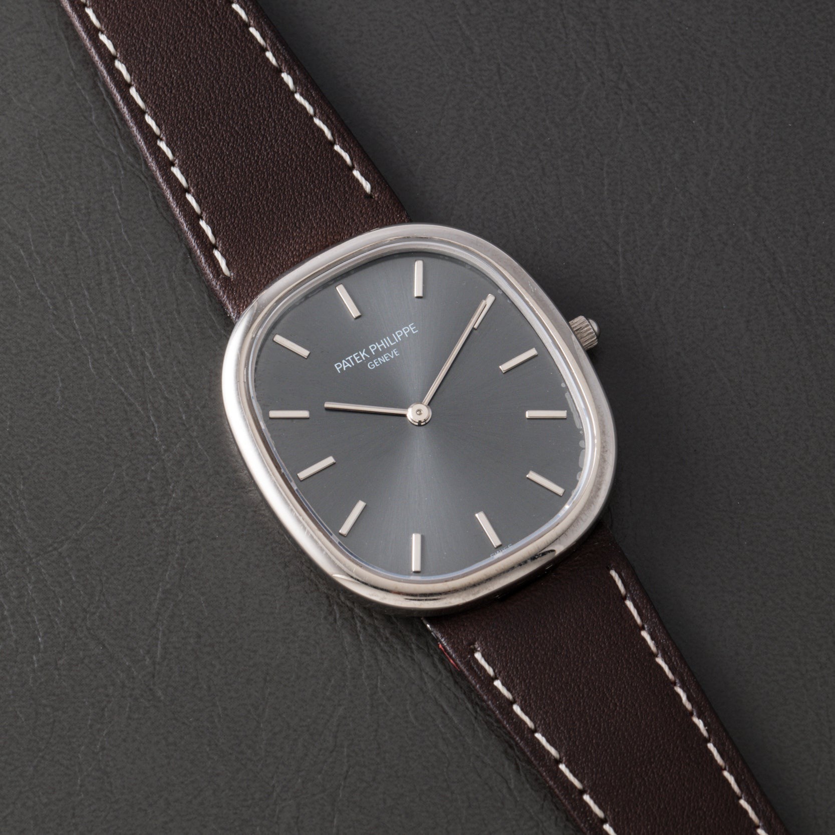 A Patek Philippe Ellipse watch with a silver case, gray face, and brown leather strap displayed on a black surface.