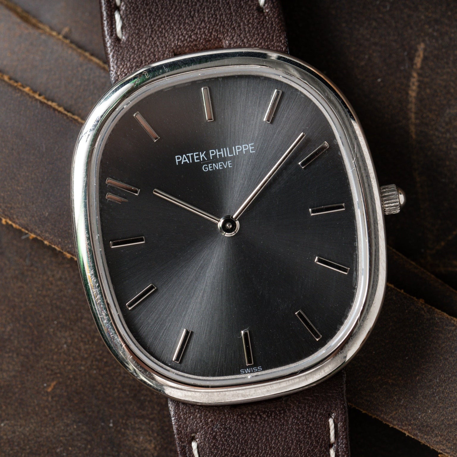 Close-up of a Patek Philippe Ellipse wristwatch featuring a black face and brown leather strap, displayed on a dark leather surface.