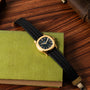 A Patek Philippe Aquanaut gold wristwatch with a black strap rests on a closed green book atop a wooden surface.