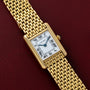 The Cartier Tank Louis wristwatch features a rectangular face with Roman numeral markers, set against a red leather surface.