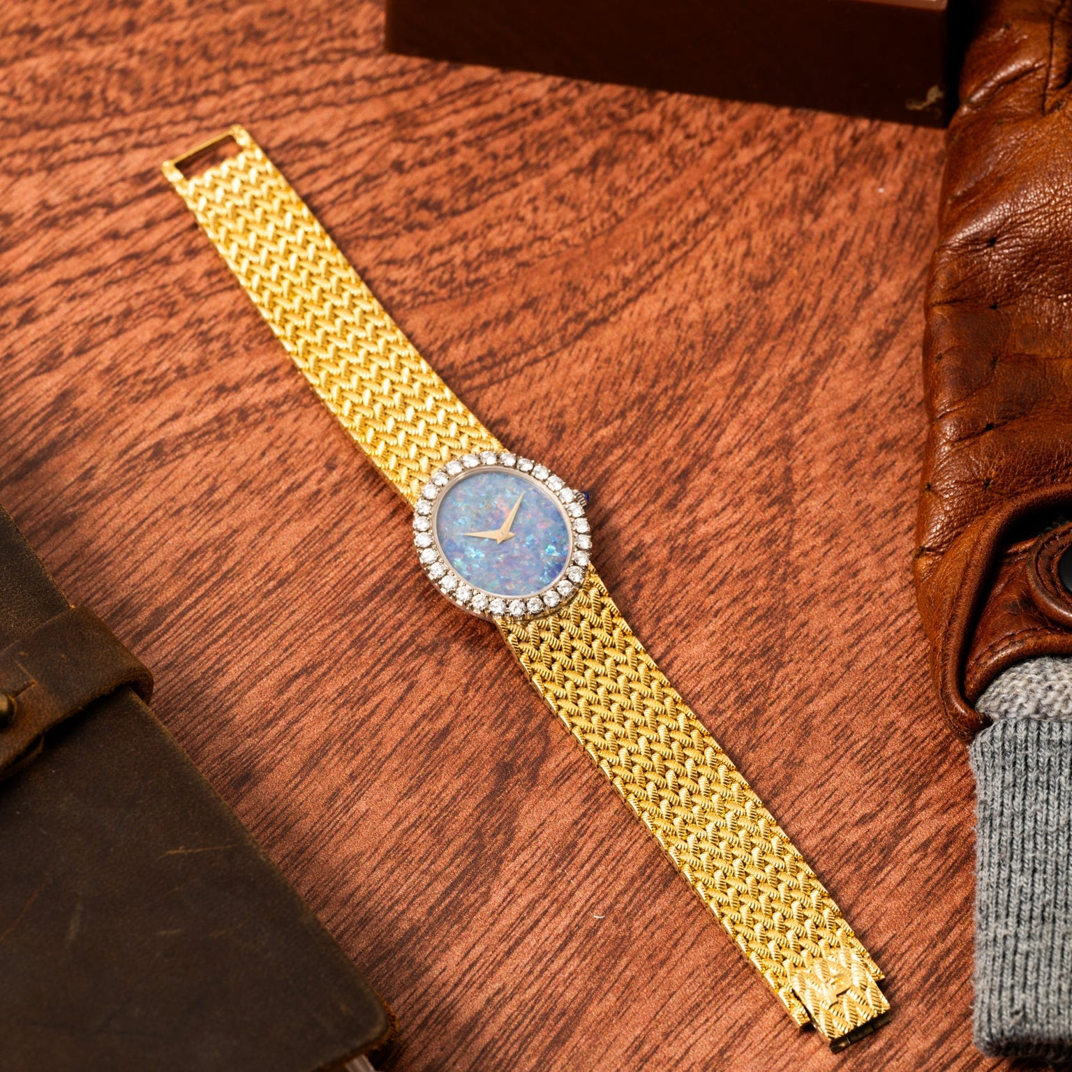The Piaget Opal Yellow Gold Dress Watch, featuring a blue face and diamond bezel, rests on a wooden surface near leather accessories.