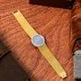 The Piaget Opal Yellow Gold Dress Watch, featuring a blue face and diamond bezel, rests on a wooden surface near leather accessories.