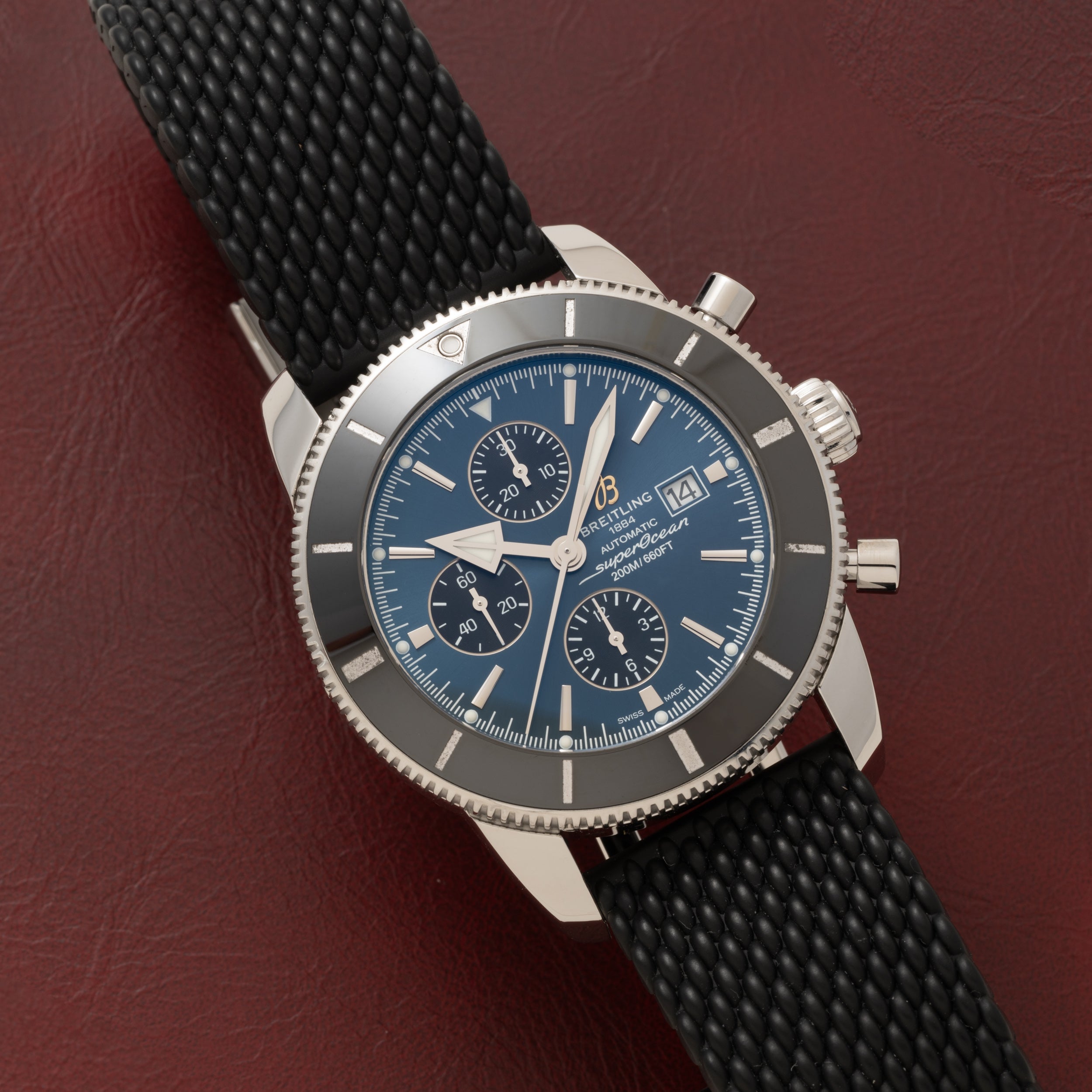 A silver Breitling SuperOcean B01 Chronograph 44 with a blue dial and black strap displayed on a textured dark red surface.