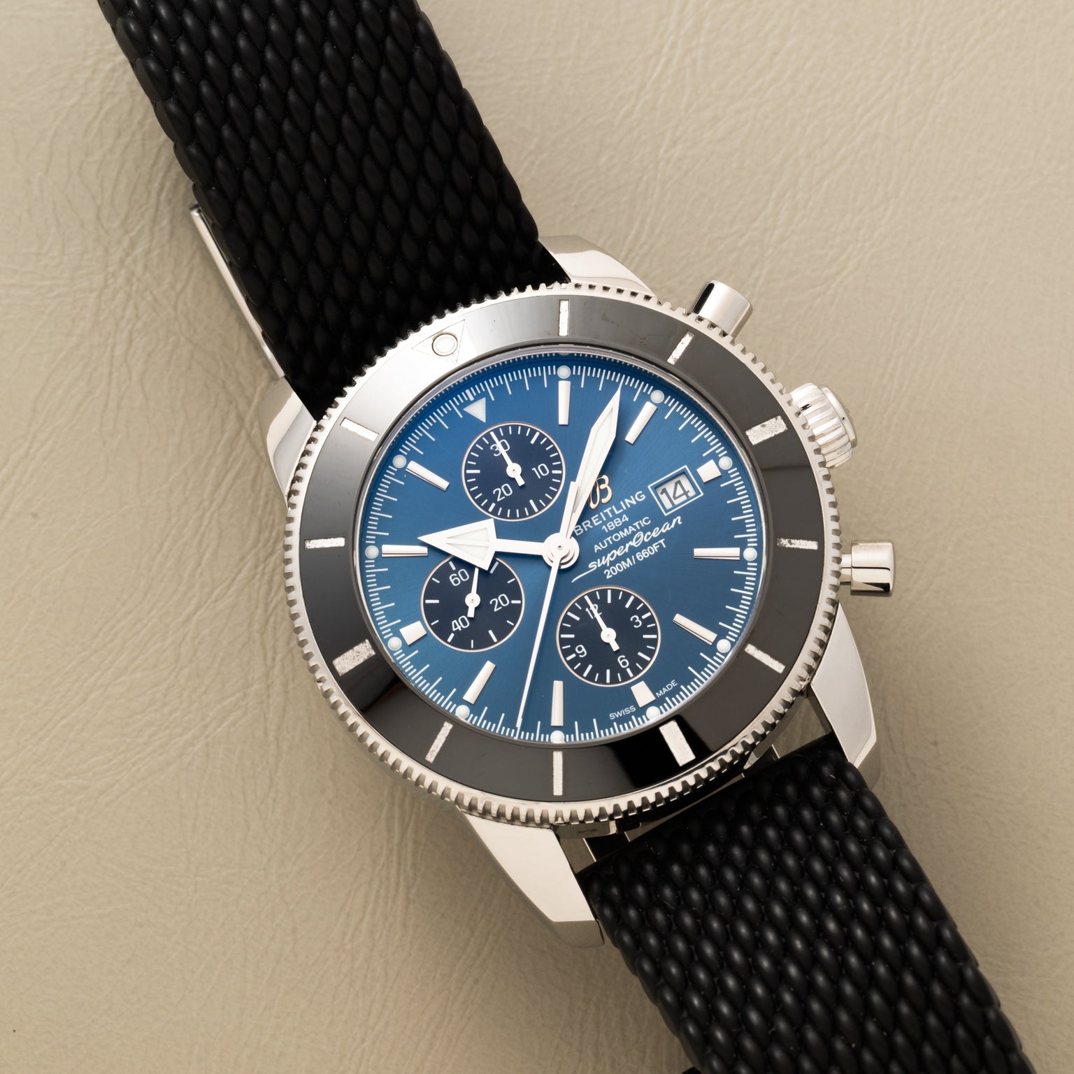 Breitling SuperOcean B01 Chronograph 44: a silver watch with a blue dial, black textured strap, and three subdials displayed on a beige surface.