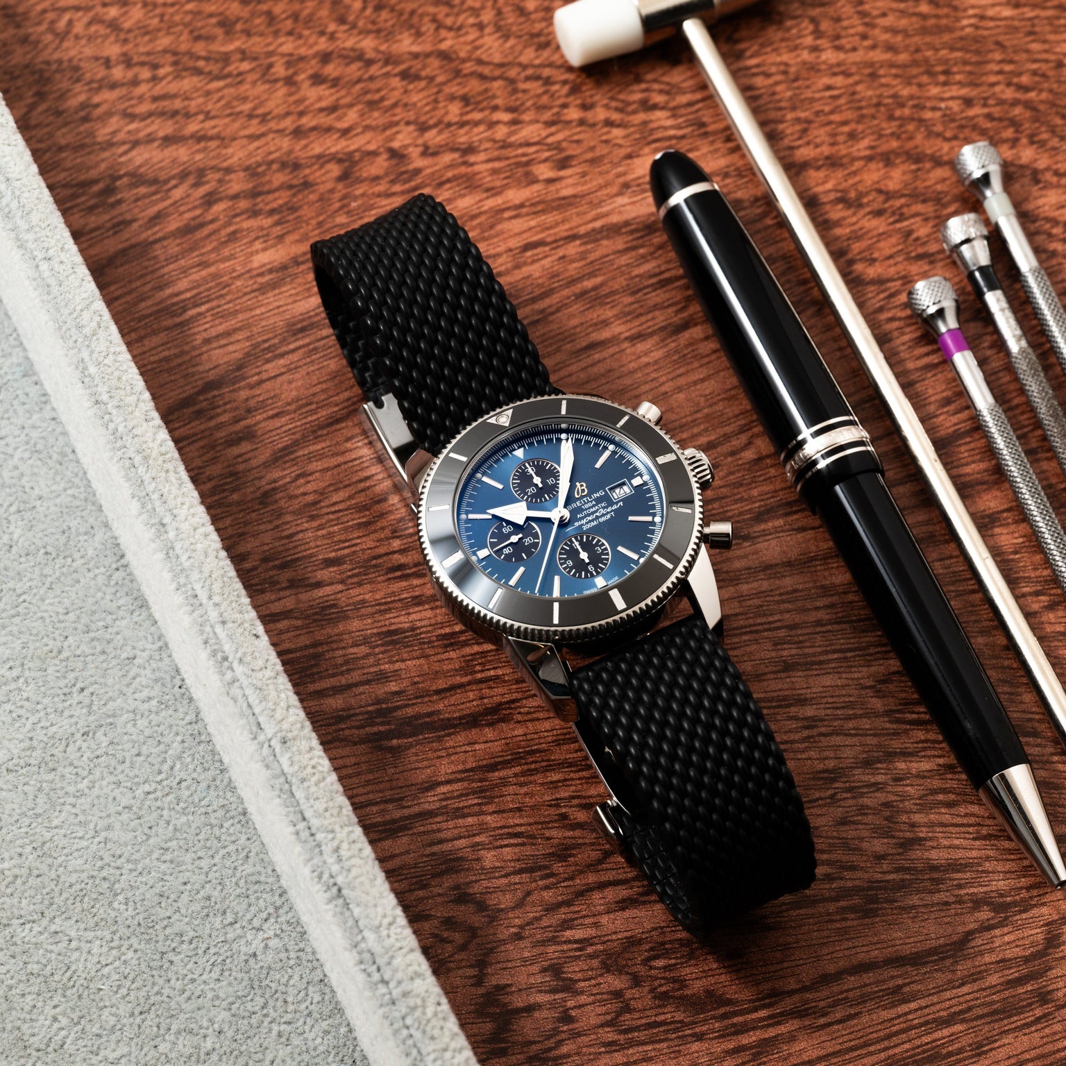A Breitling SuperOcean B01 Chronograph 44 wristwatch with a black strap, paired with a black pen and metal tools on a wooden surface.