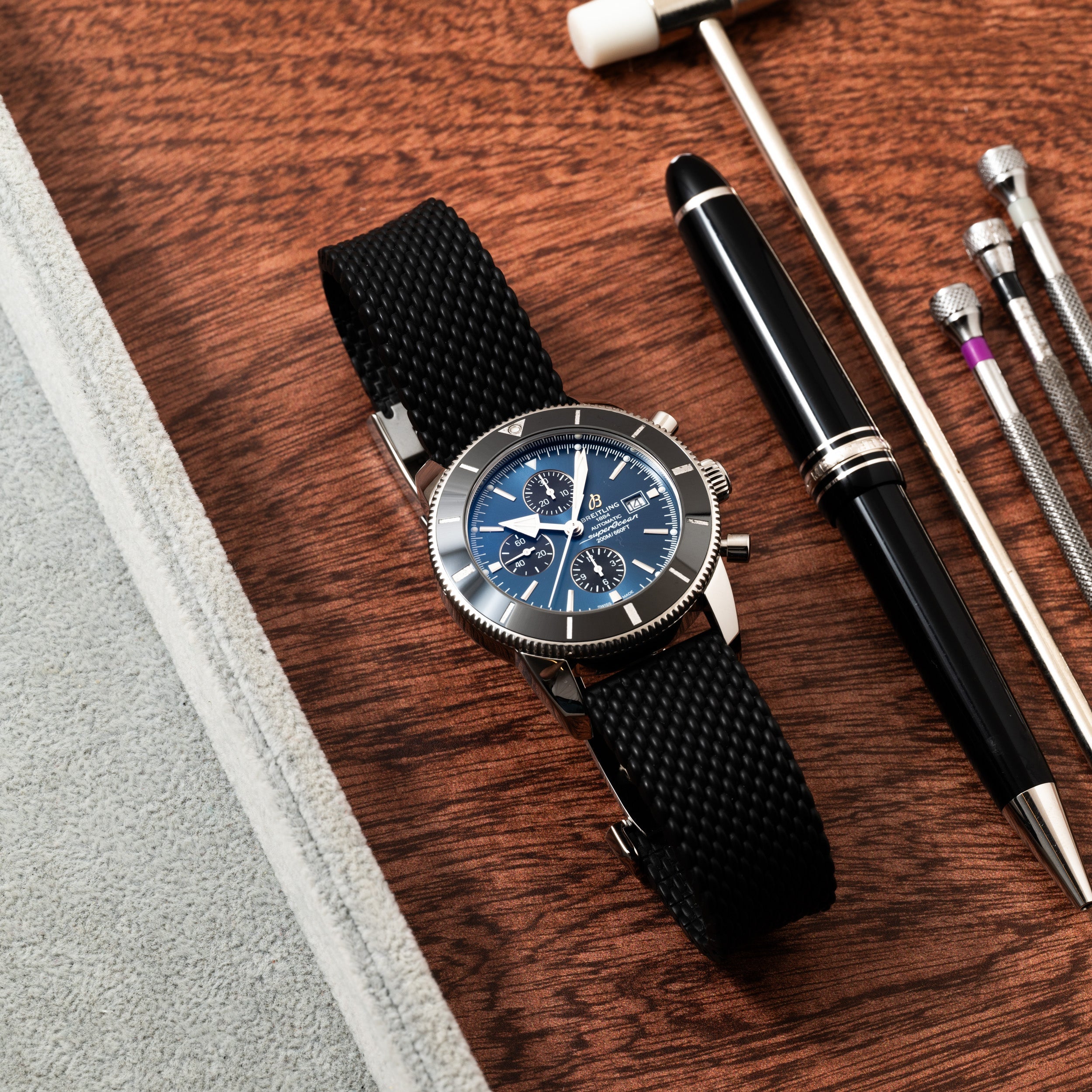 A Breitling SuperOcean B01 Chronograph 44 wristwatch with a black strap, paired with a black pen and metal tools on a wooden surface.