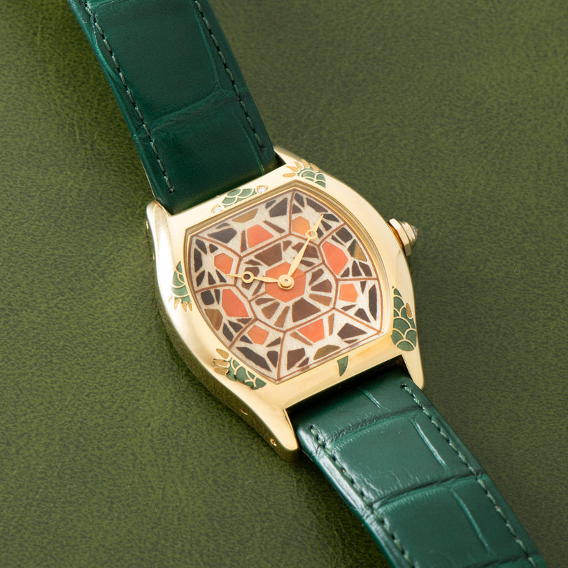 The Cartier Tortue XL Turtle Limited Edition wristwatch features a geometric turtle shell-patterned face and a green leather strap, displayed on a green surface.