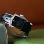 Close-up of a Patek Philippe Calatrava Diamond Bezel watch, featuring a black leather strap with a silver buckle, displayed on green and beige surfaces.