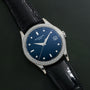 The Patek Philippe Calatrava Diamond Bezel features a blue dial, diamond-set bezel, and black leather strap, elegantly displayed on a dark surface.