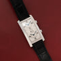 A rectangular Cartier Tank Cintrée CPCP Dual-Time wristwatch with two dials and a black leather strap is displayed on a red background.