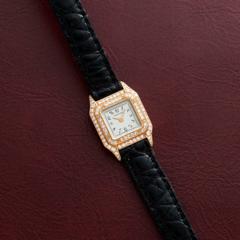 The Cartier Panthère gold watch features diamonds and a black leather strap, displayed on a textured dark red background.