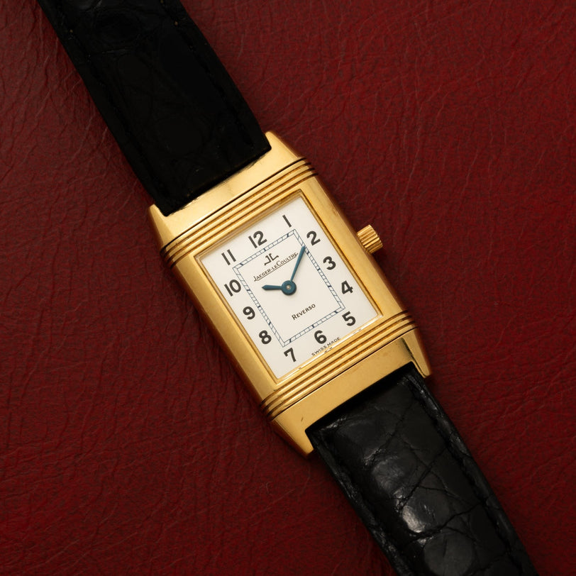 A Jaeger-LeCoultre Reverso Classique gold rectangular wristwatch with a black leather strap, displayed on a textured red surface.