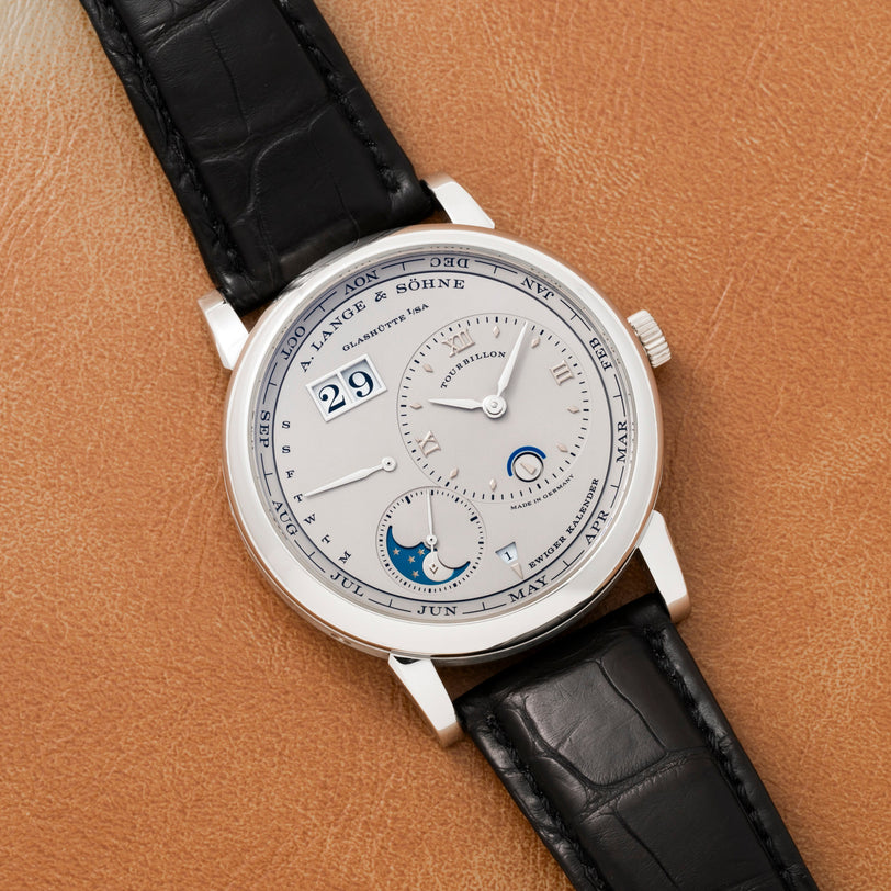 The Lange & Sohne Lange 1 Tourbillon Perpetual Calendar is a luxury wristwatch with a black leather strap on a tan surface, featuring multiple dials and date indicators.