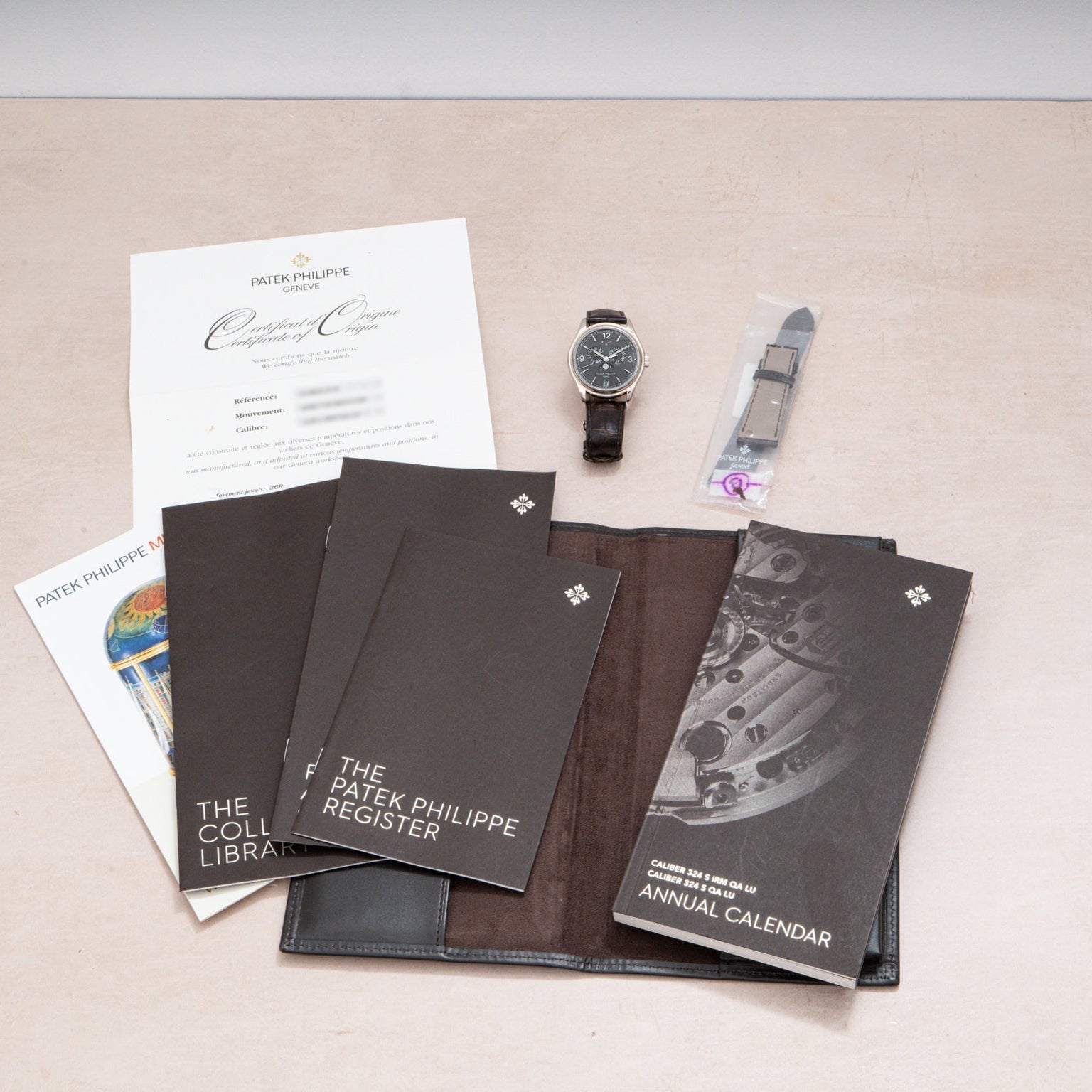 A Patek Philippe Annual Calendar watch with documents, manuals, certificate, strap, and a black leather folder on a table.