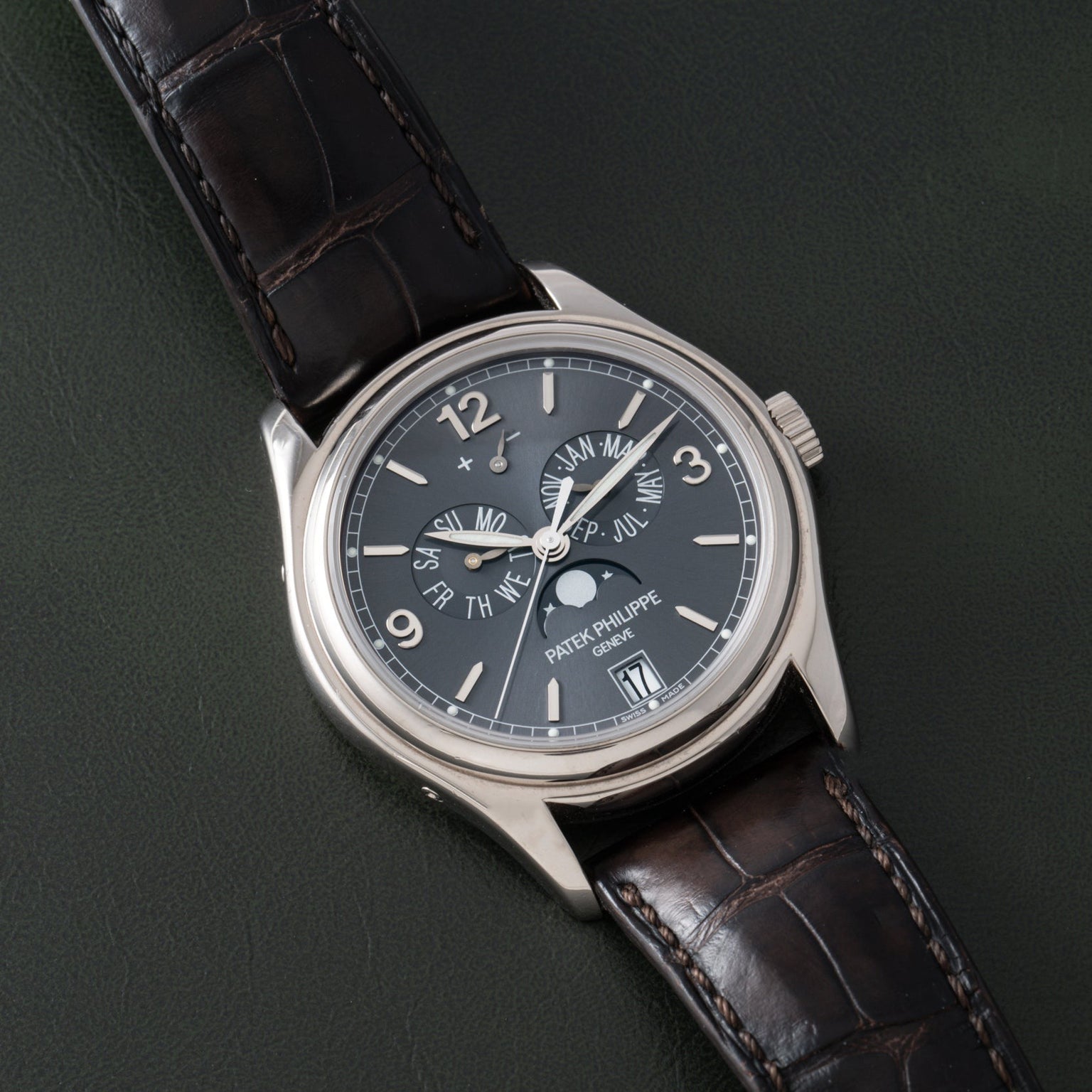 The Patek Philippe Annual Calendar, a luxury wristwatch with a dark face and leather strap, is displayed on a dark surface.