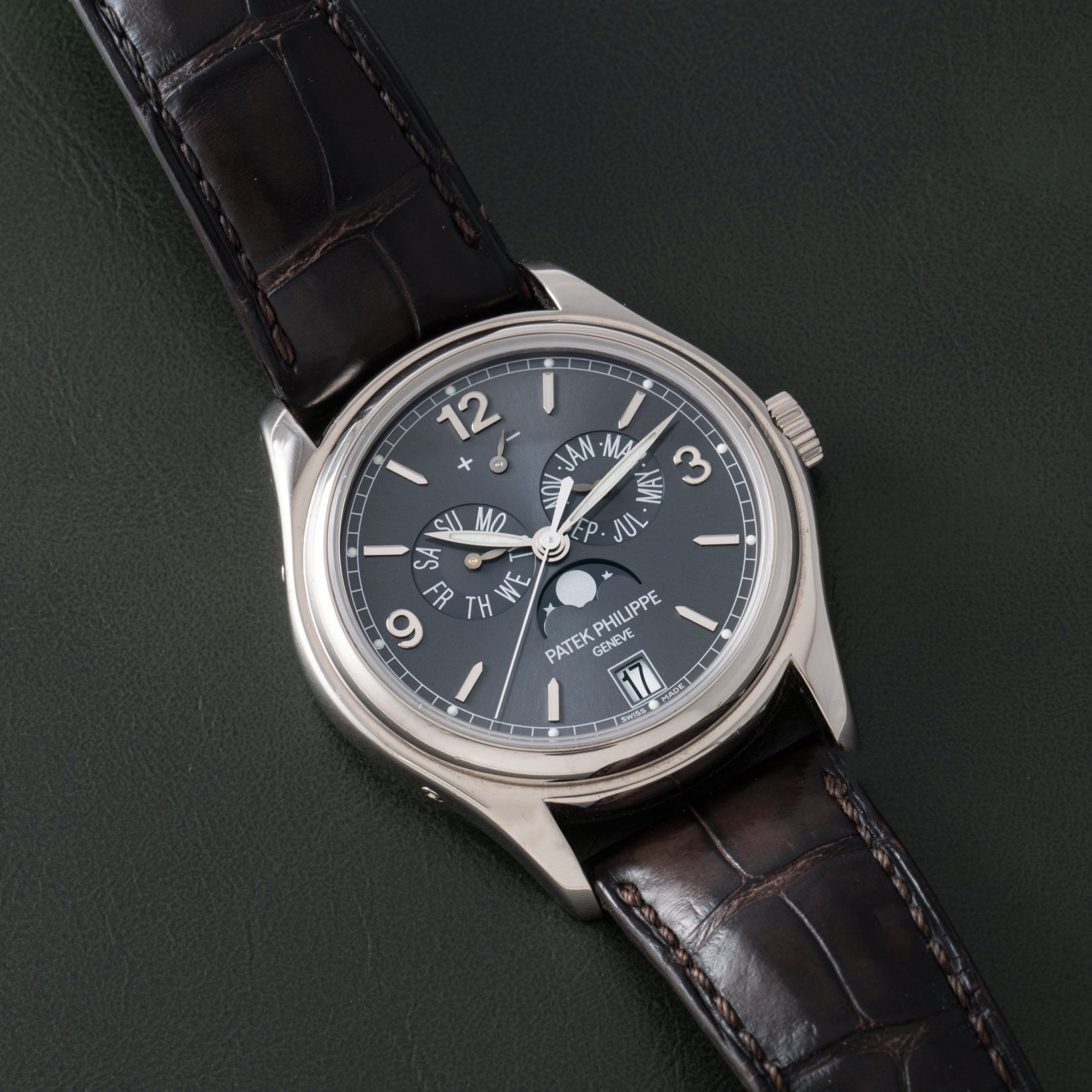 The Patek Philippe Annual Calendar, a luxury wristwatch with a dark face and leather strap, is displayed on a dark surface.