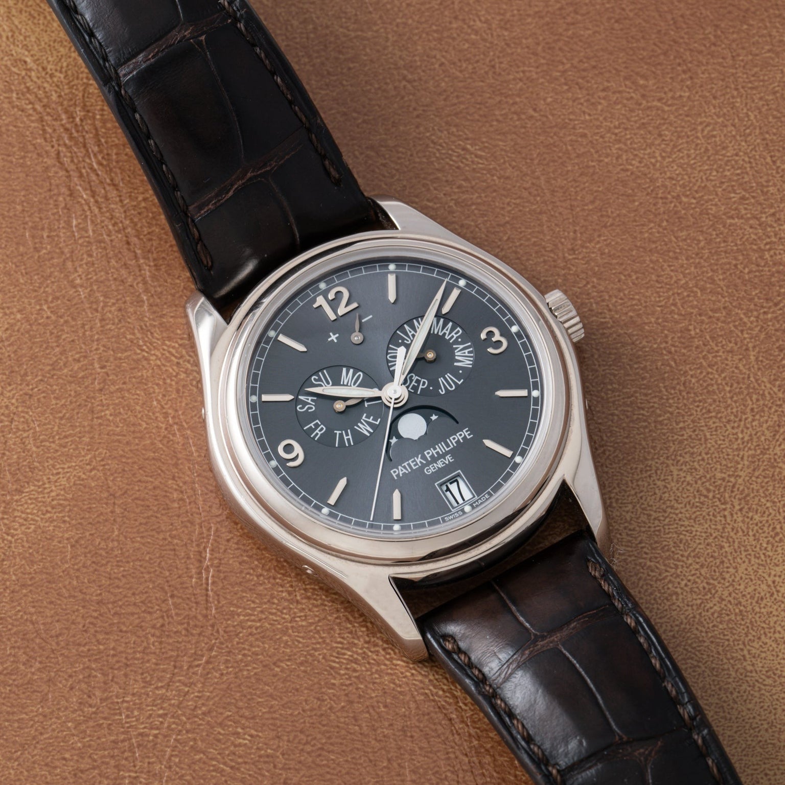 The Patek Philippe Annual Calendar wristwatch features a black dial and a brown leather strap, displayed on a matching brown leather surface.