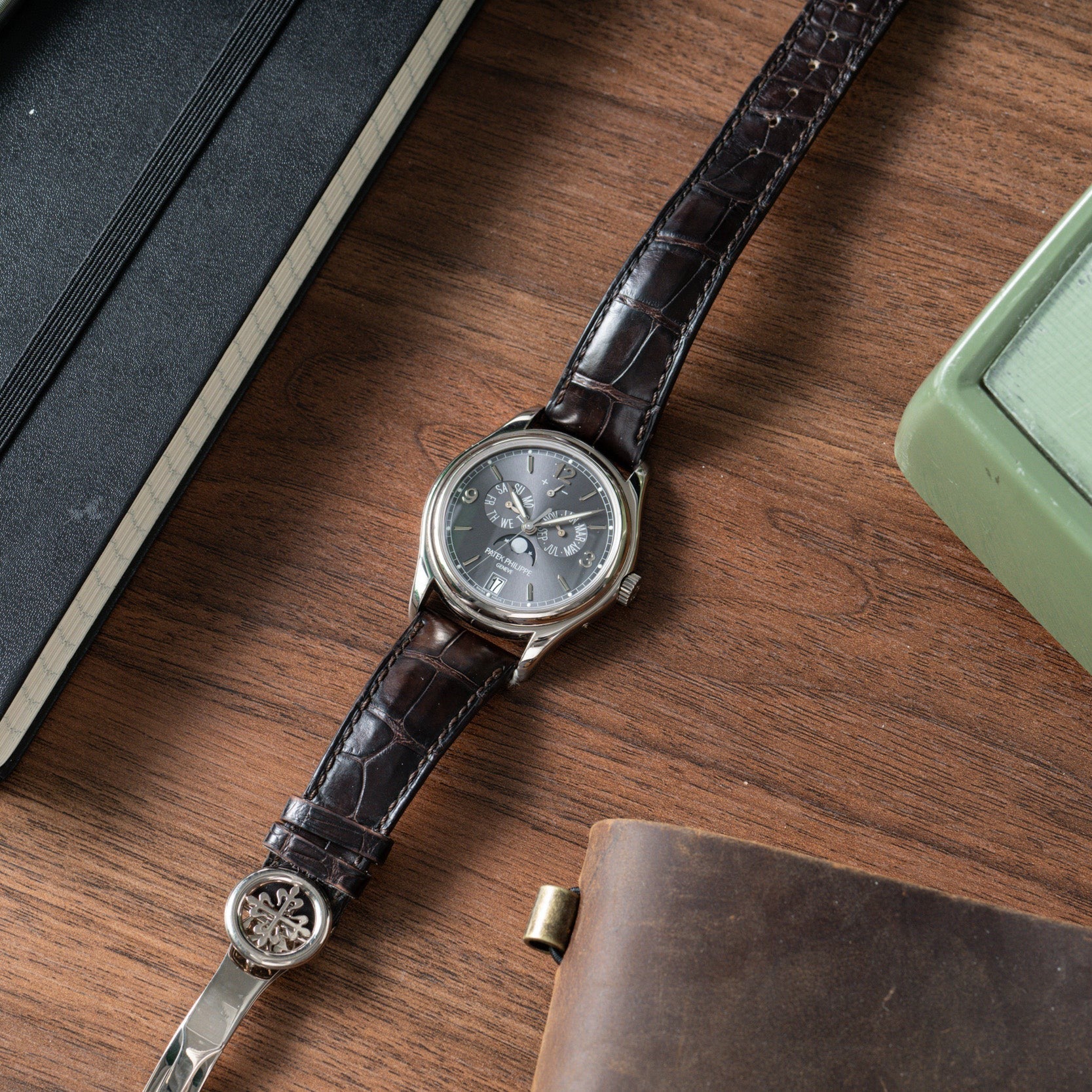 A Patek Philippe Annual Calendar wristwatch with a brown leather strap rests on a wooden desk next to books and a green device.