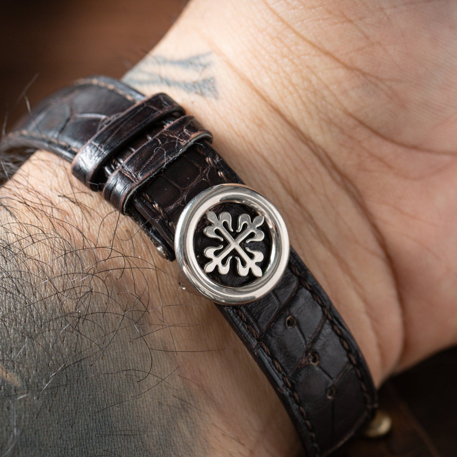 A close-up of Patek Philippes Annual Calendar watch featuring a brown leather strap and silver clasp on a tattooed wrist.