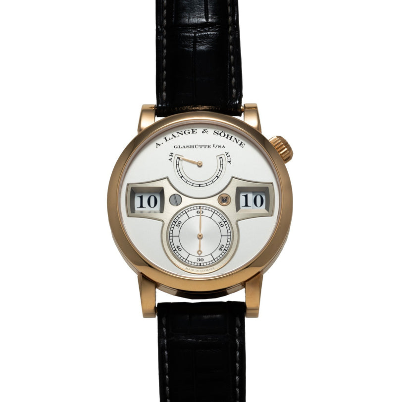 The Lange & Sohne Zeitwerk is a gold wristwatch featuring a white dial with digital hour and minute display and a black leather strap.