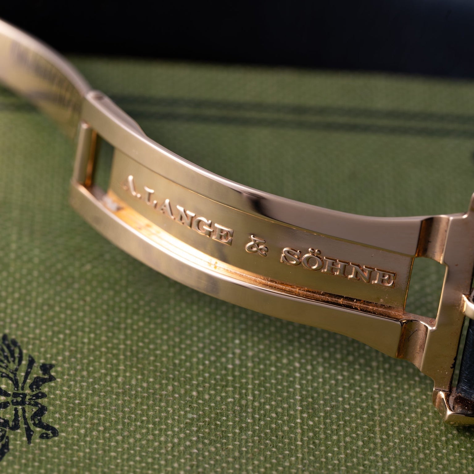 Close-up of a gold Zeitwerk watch clasp engraved with Lange & Sohne, set on a textured green surface.