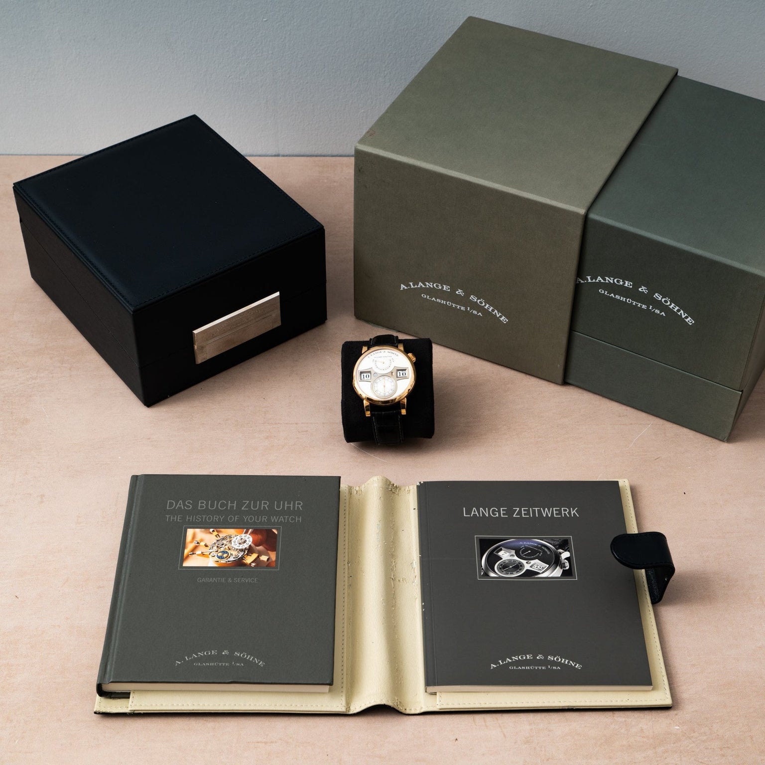 A Lange & Sohne Zeitwerk luxury watch with a black case is displayed on a beige surface alongside two gray booklets.