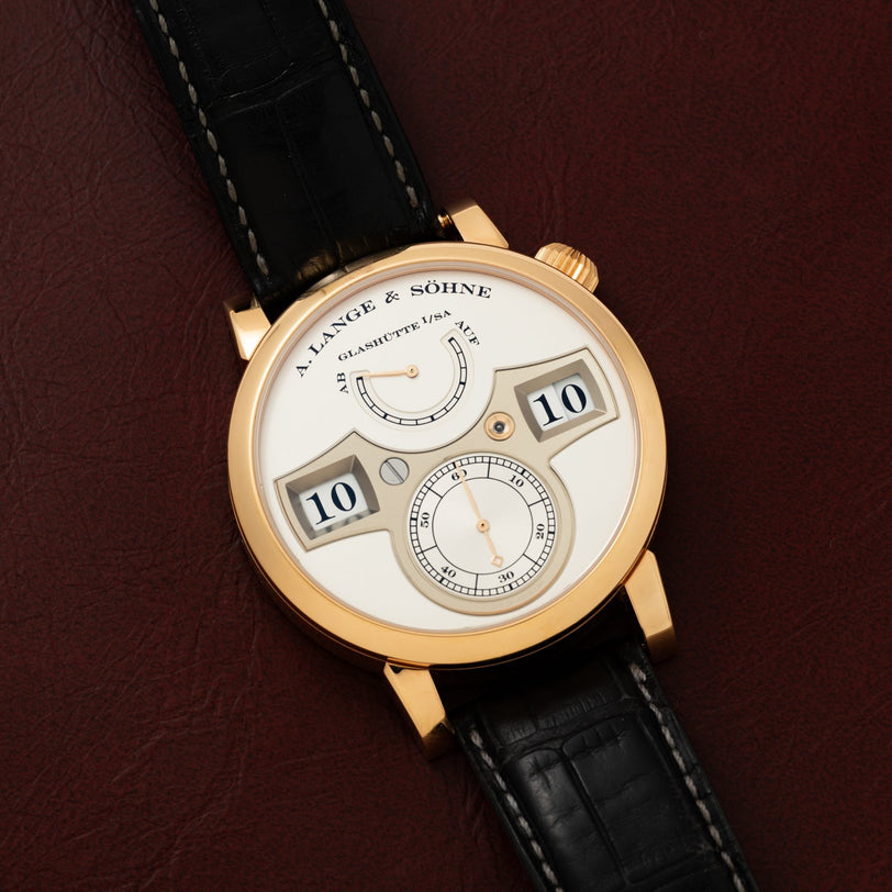 A Lange & Sohne Zeitwerk wristwatch featuring a gold case and black leather strap, displayed on a brown leather surface.