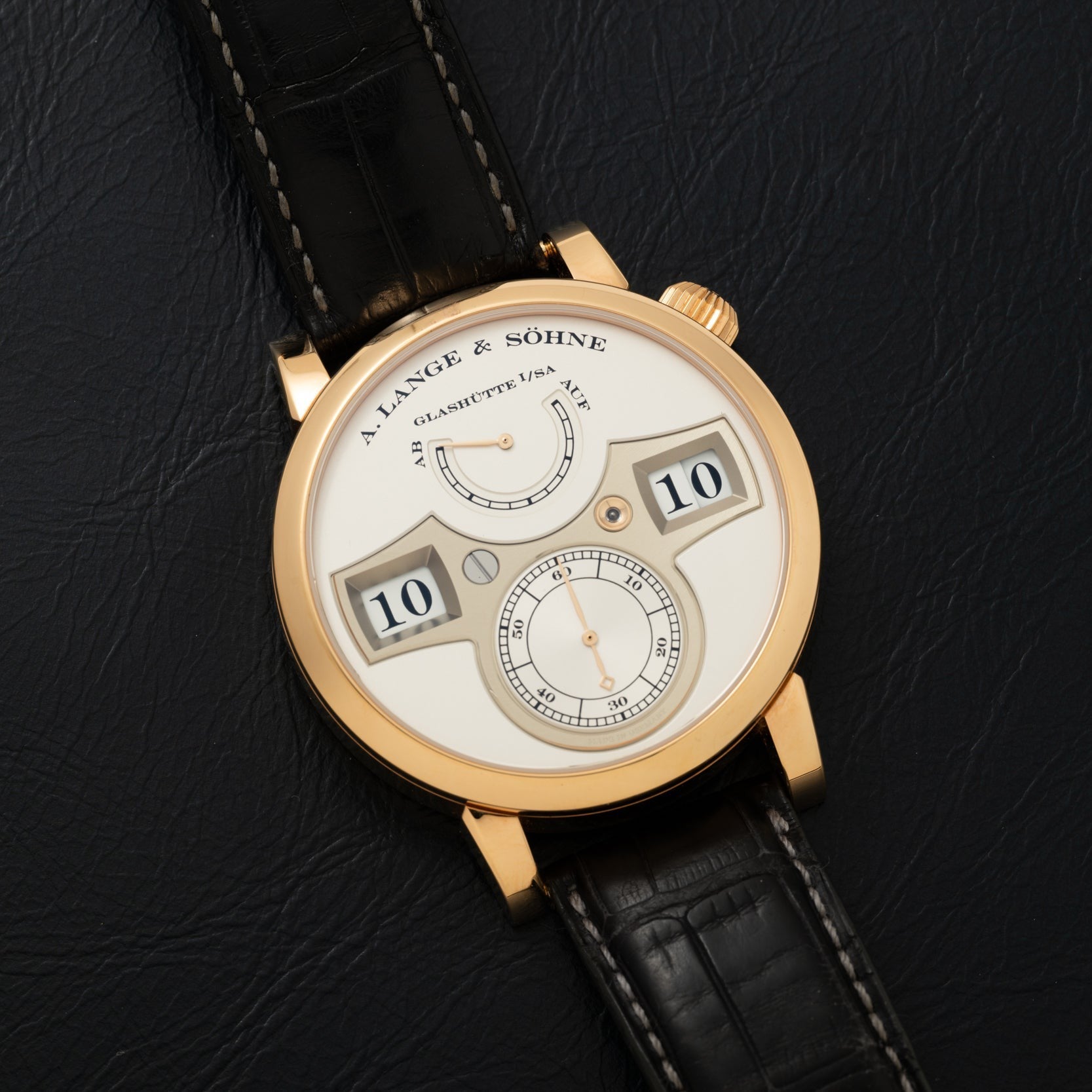 The Lange & Sohne Zeitwerk watch features a digital time display and black leather strap, shown on a black surface.