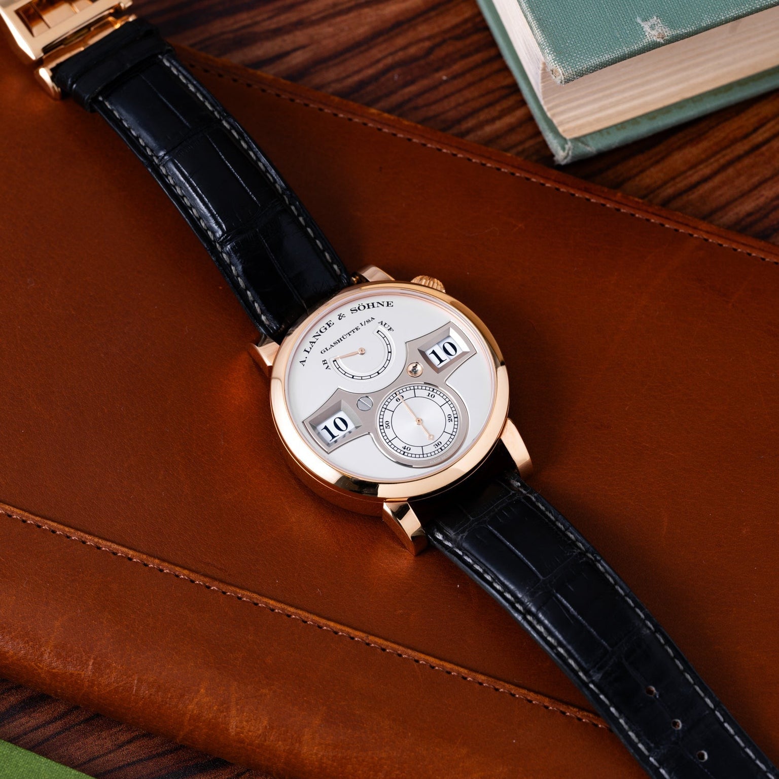 The Lange & Sohne Zeitwerk luxury wristwatch with a black leather strap is displayed on a brown leather case beside a stack of books.