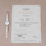 A white Patek Philippe Calatrava wristwatch rests next to its certificate of origin on a beige surface.