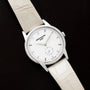 A Patek Philippe Calatrava wristwatch featuring a diamond bezel and white leather strap, displayed on a black surface.