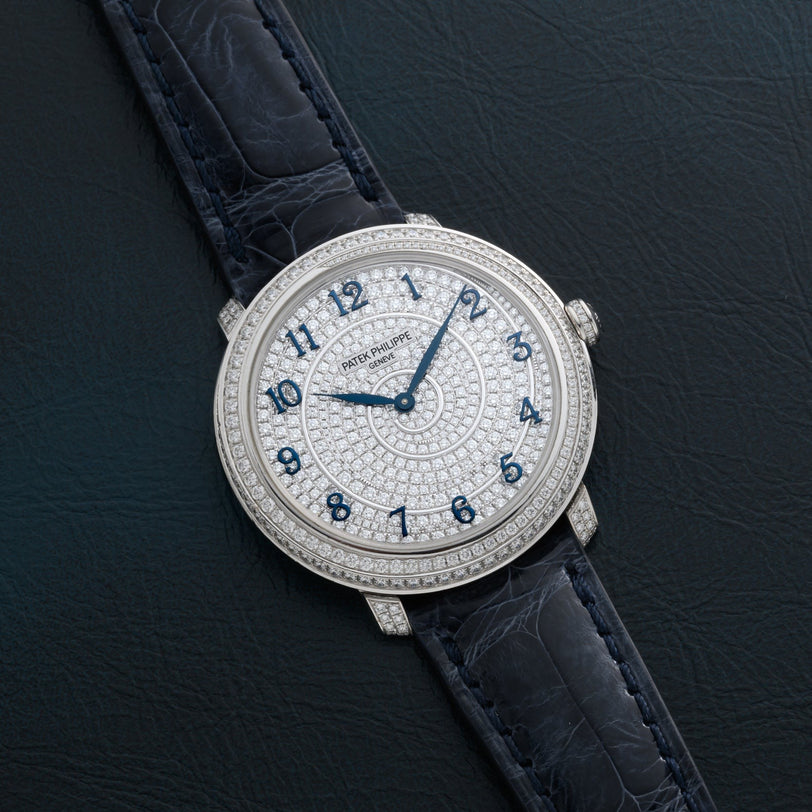 A Patek Philippe Calatrava watch, diamond-encrusted with blue numerals and a dark leather strap, displayed on a black surface.