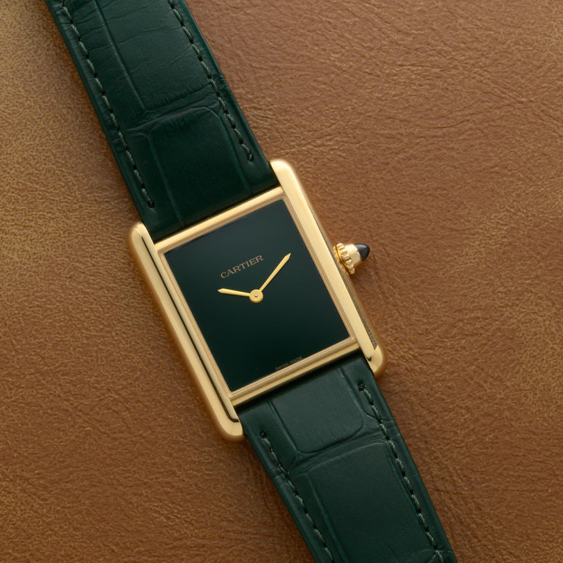A Cartier Tank Louis watch with a gold case, green face, and green leather strap sits on a brown leather surface.