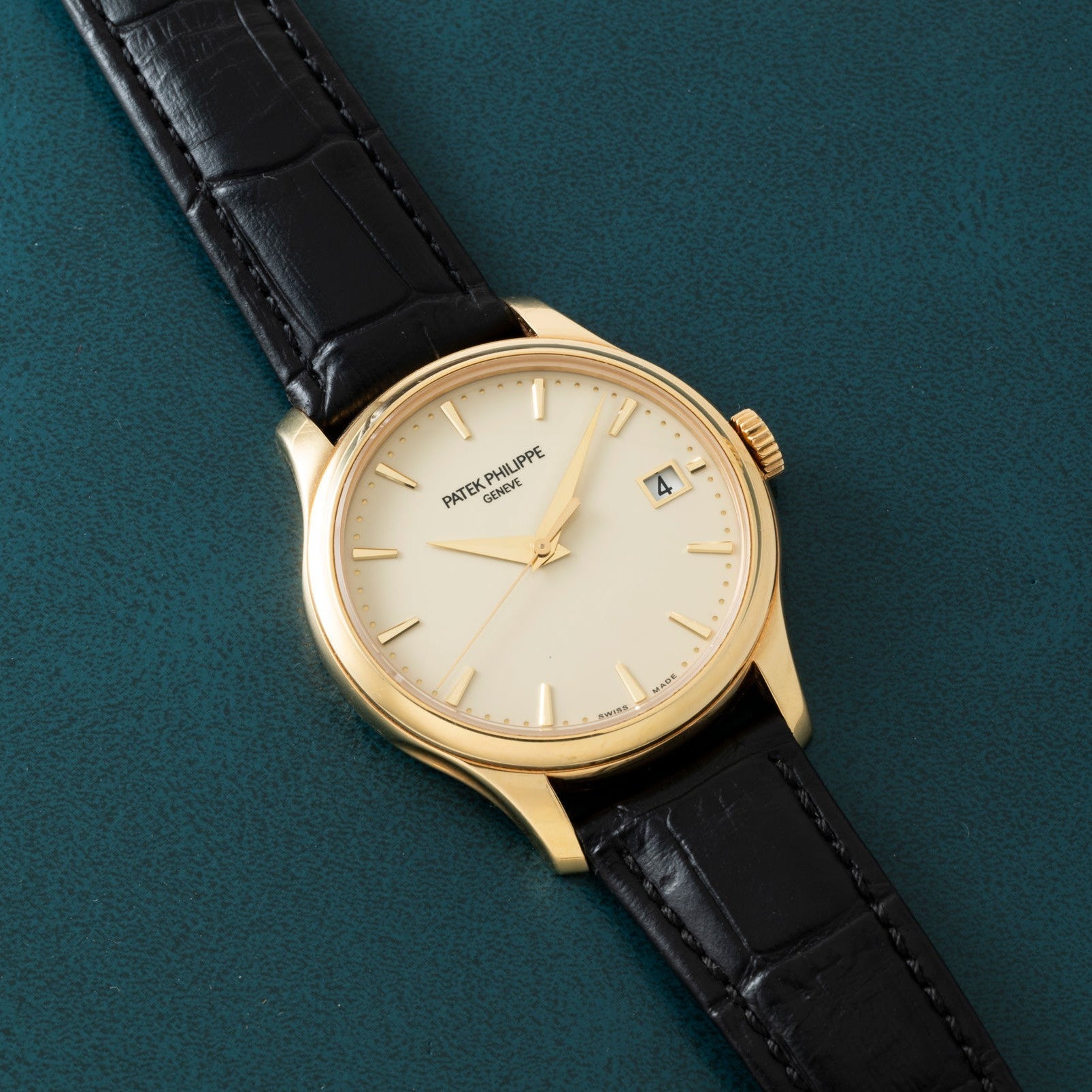 The Patek Philippe Calatrava, featuring a gold case, black leather strap, and date display, is displayed on a green textured surface.