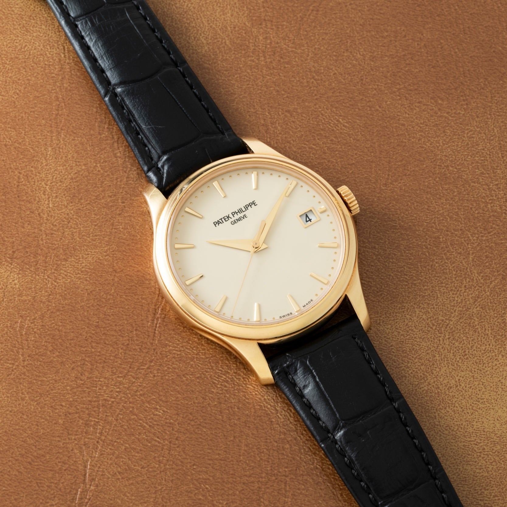 A Patek Philippe Calatrava gold wristwatch with a black leather strap rests on a brown leather surface.