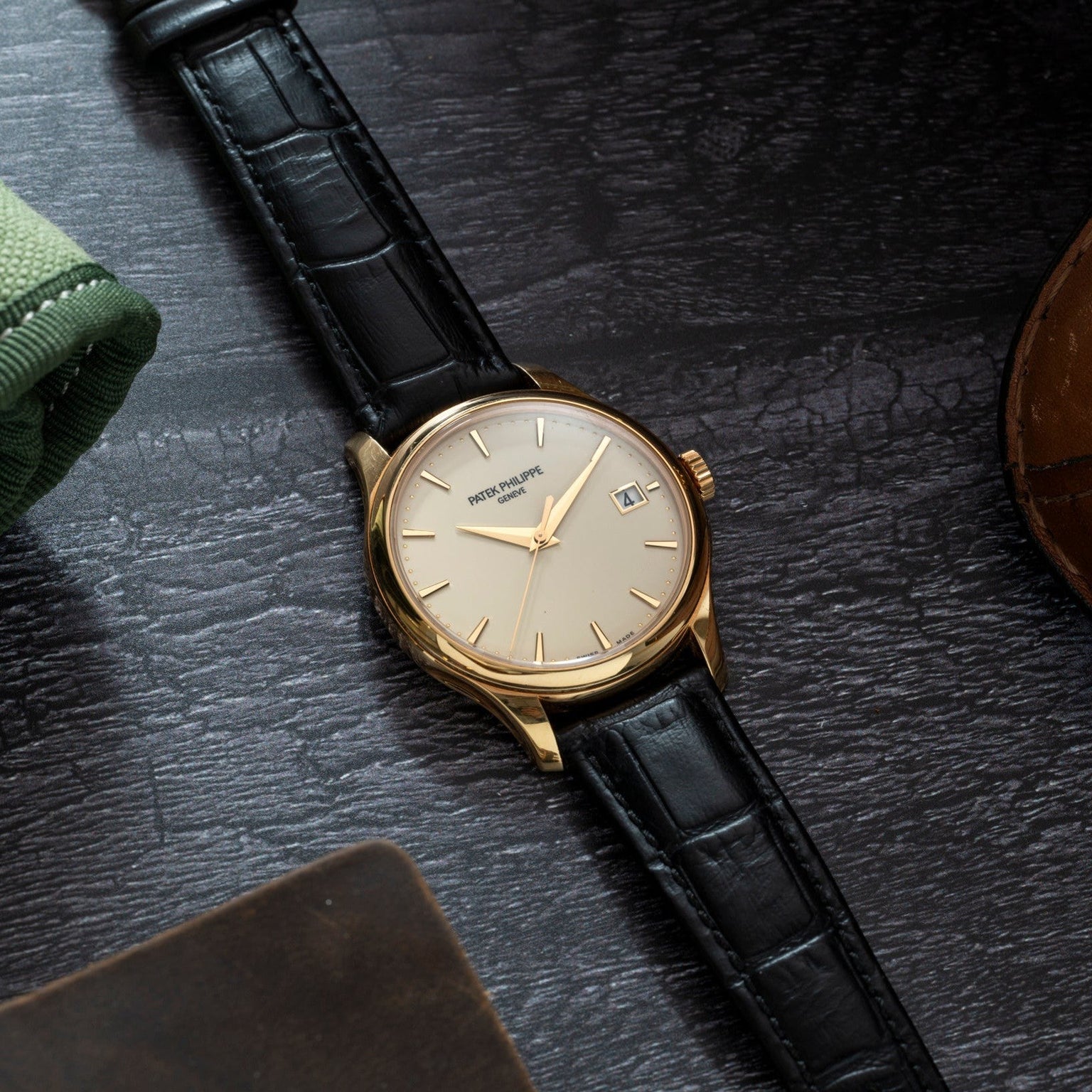 The Patek Philippe Calatrava gold wristwatch with a black leather strap rests on a dark wooden surface beside green and brown fabric accessories.
