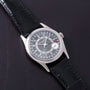 A Patek Philippe Calatrava wristwatch in silver with a black leather strap, displayed on a dark leather surface.