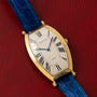 Cartier Tonneau XL wristwatch with Roman numerals and blue leather strap, displayed on a red textured surface.